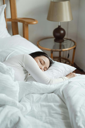 Woman sleeping peacefully in bed at home. Woman is wearing white and sleeping in a fluffy and comfortable bed set. The decor of house has warm wood tones.